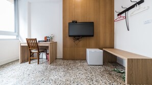 Ein kleines Zimmer mit Schreibtisch aus Holz, einem Stuhl, einem kleinen weißen Kühlschrank und einem Wandfernseher.