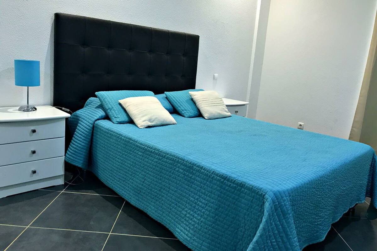 A neatly made bed with a blue quilt and pillows, a bedside lamp, and a white nightstand with drawers.