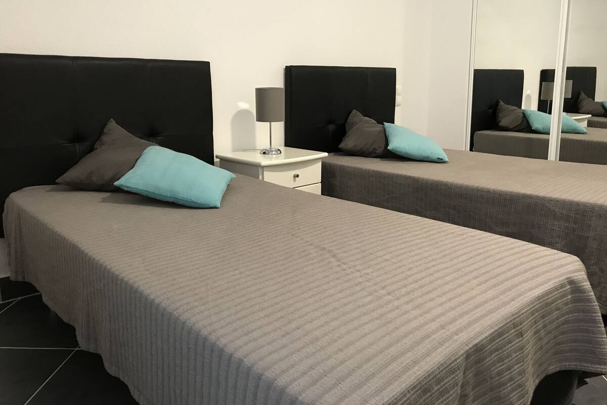 A hotel room with two beds, each with a grey bedspread and a single pillow. There is a white bedside table with a lamp and a small drawer. The room has a large mirror on one wall.