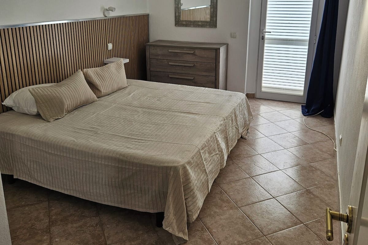 A bedroom with a bed, a dresser, a mirror, an air conditioning unit, and a door with a curtain.