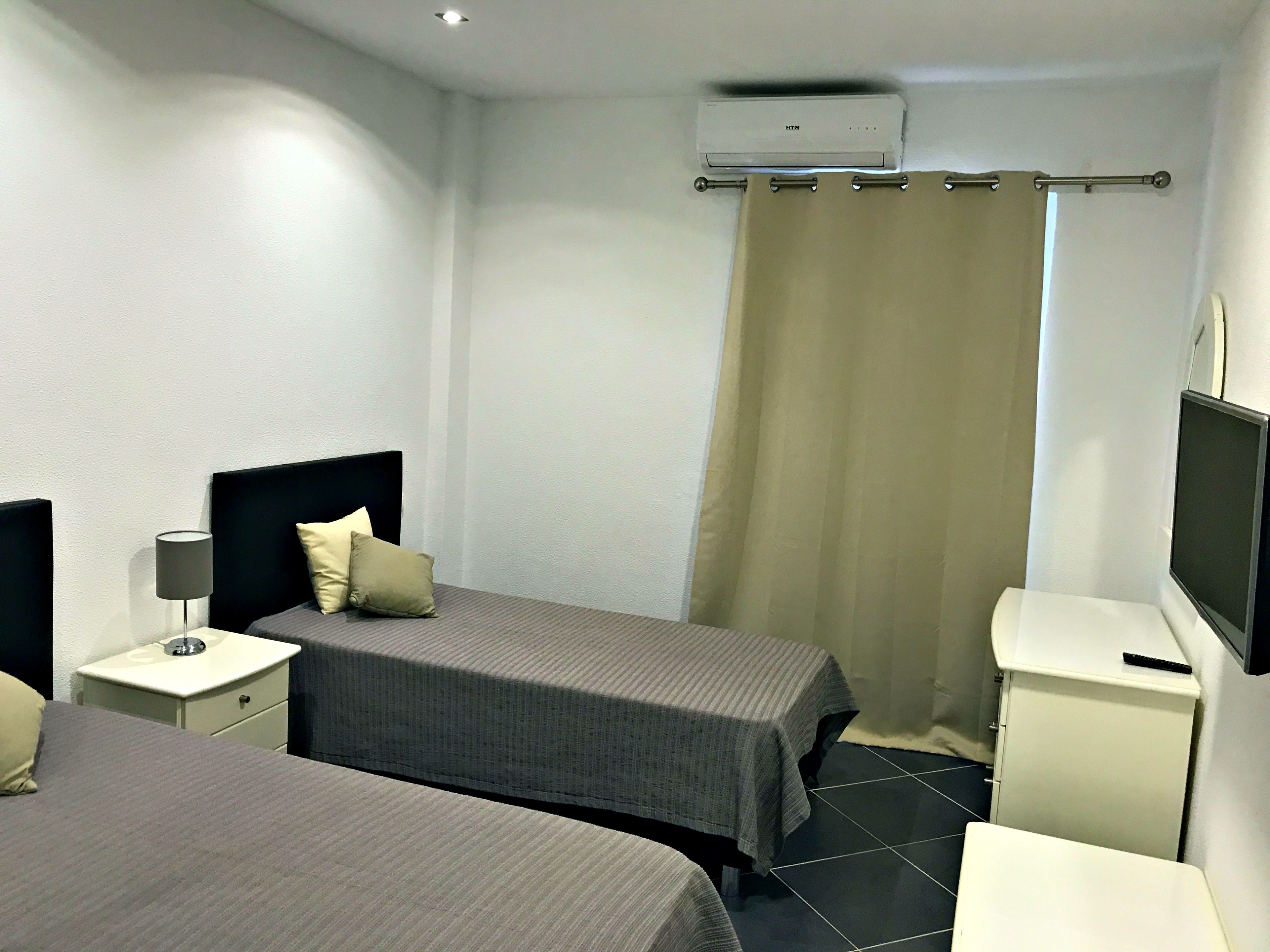 A hotel room with two beds, a bedside table with a lamp, a TV, and an air conditioning unit.