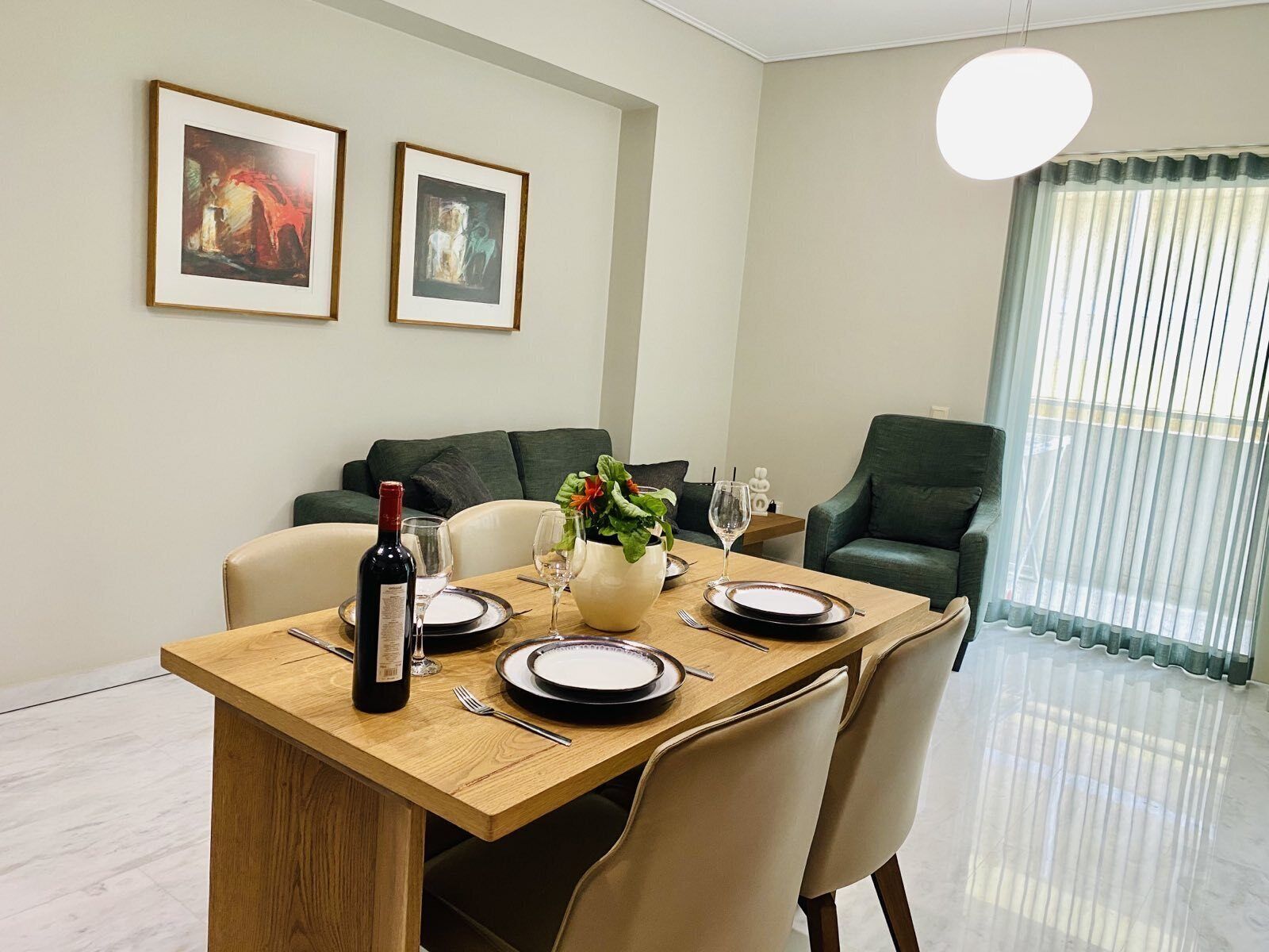 Un comedor con mesa de madera dispuesta para cuatro personas, dos cuadros en la pared, un sofá verde y vistas a través de una ventana con cortinas traslúcidas.