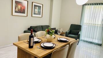 Un comedor con mesa de madera dispuesta para cuatro personas, dos cuadros en la pared, un sofá verde y vistas a través de una ventana con cortinas traslúcidas.