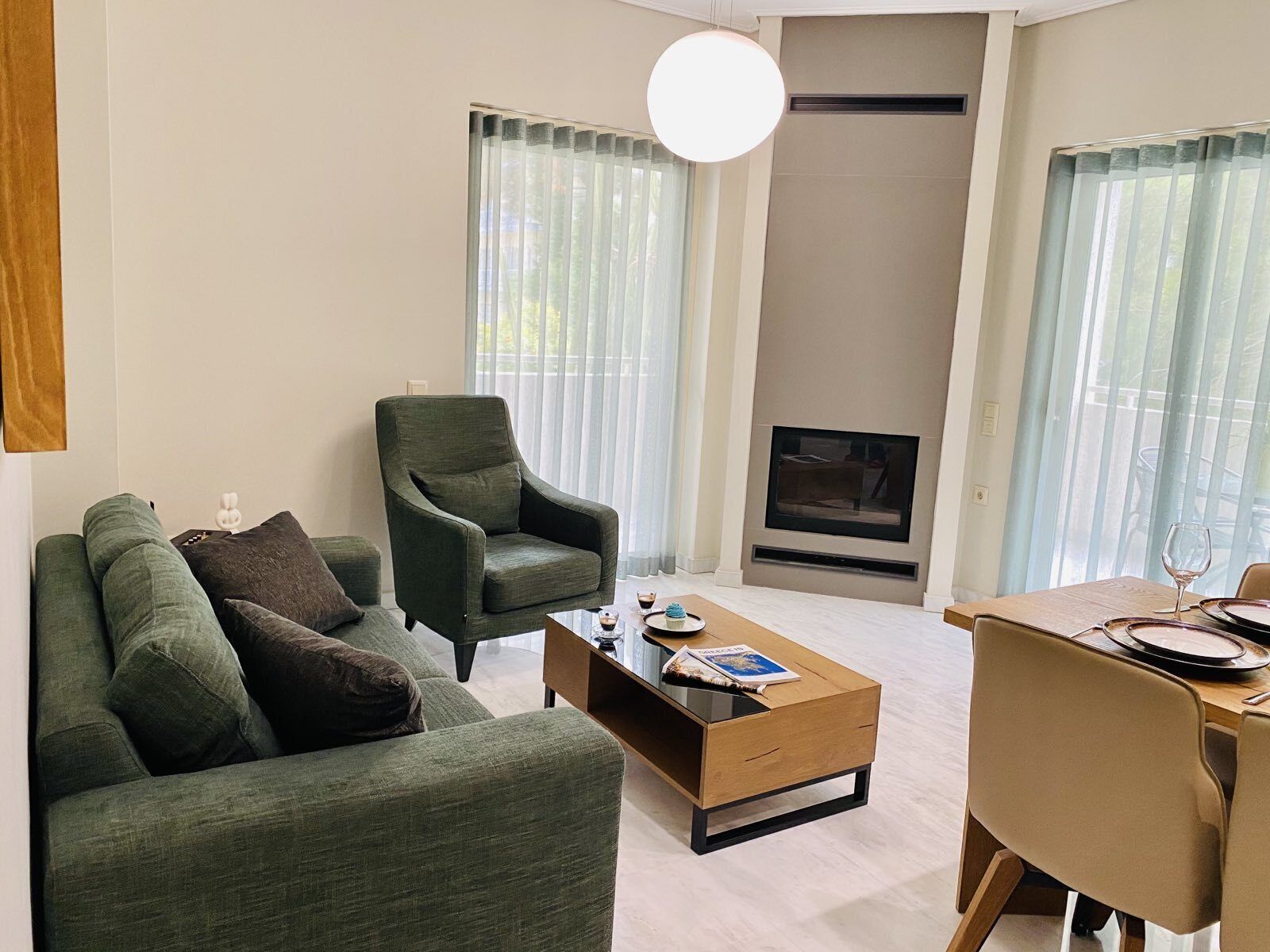 A modern living room with a green sofa, a green armchair, a wooden coffee table, and a dining table with plates and a wine glass.