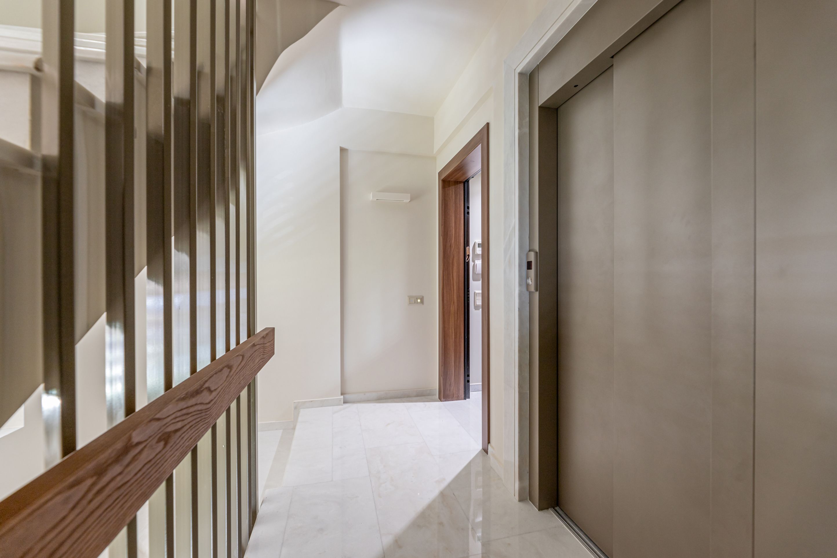 A hallway with a wooden handrail, a white tiled floor, and a closed door at the end.