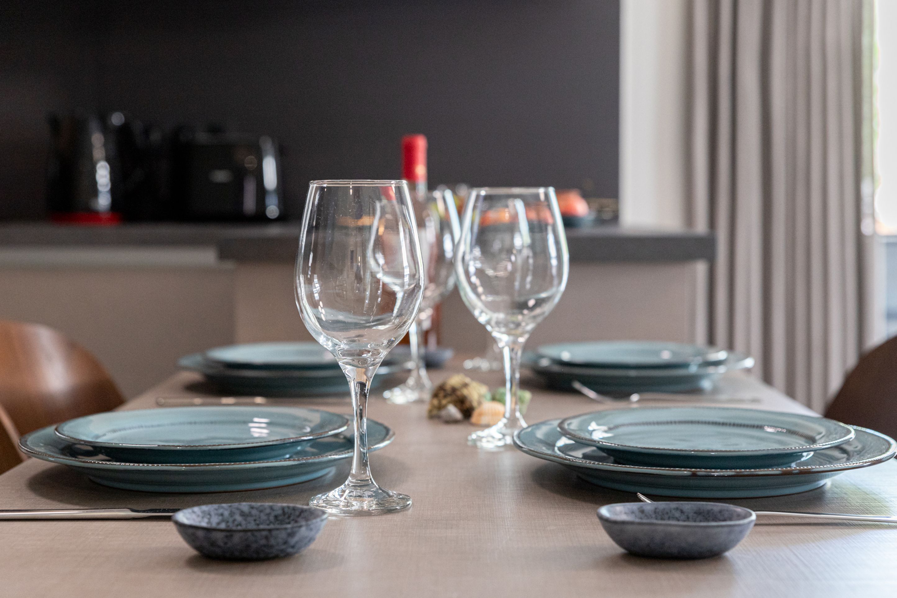 A dining table set with clear glassware and teal plates, with a kitchen in the background.