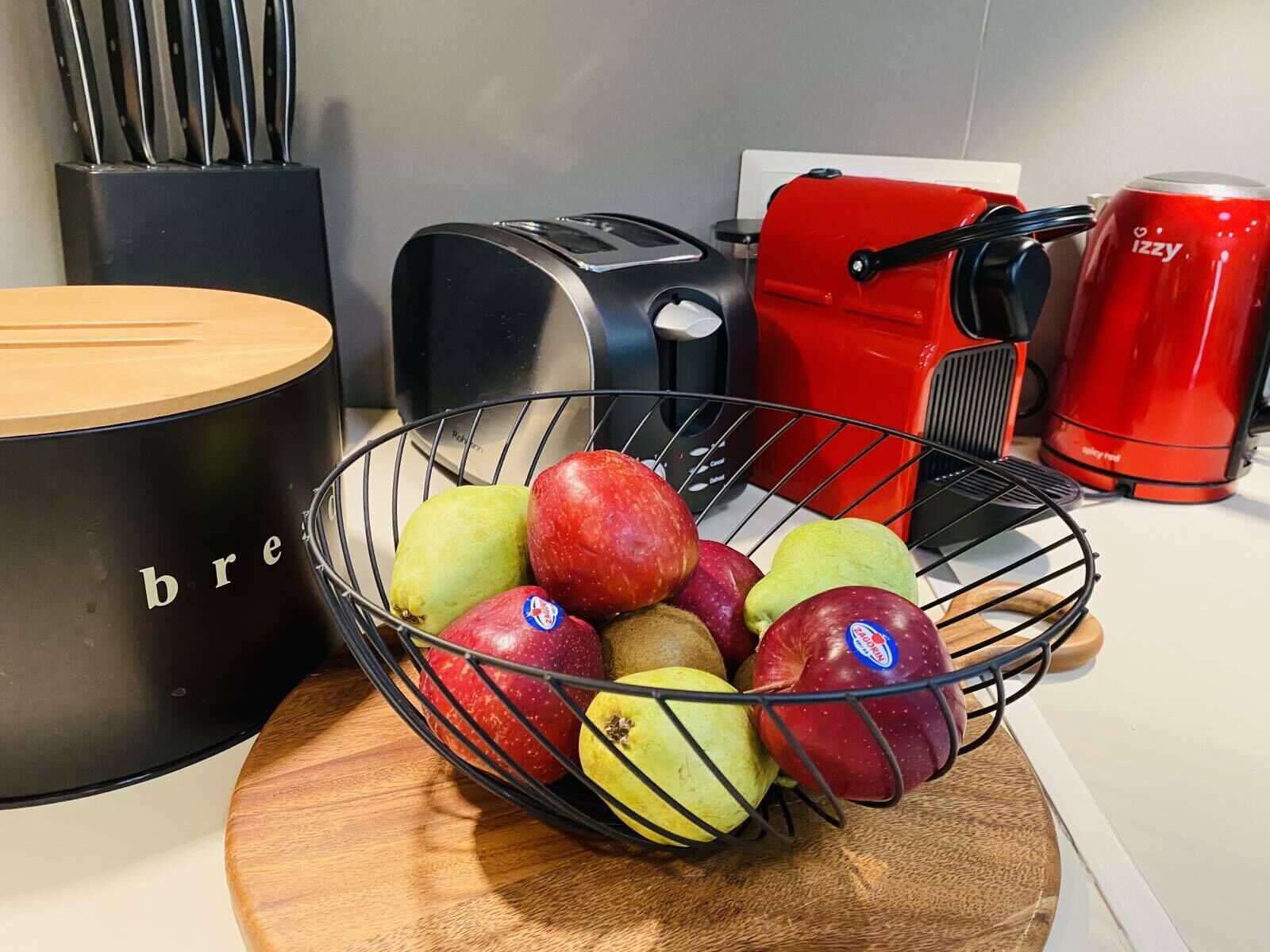 Eine Küchenarbeitsplatte mit Obstschale, gefüllt mit Äpfeln und Birnen, einem Toaster, einer roten Kaffeemaschine und einem schwarzen Behälter, auf dem das Wort 'bre' sichtbar ist.