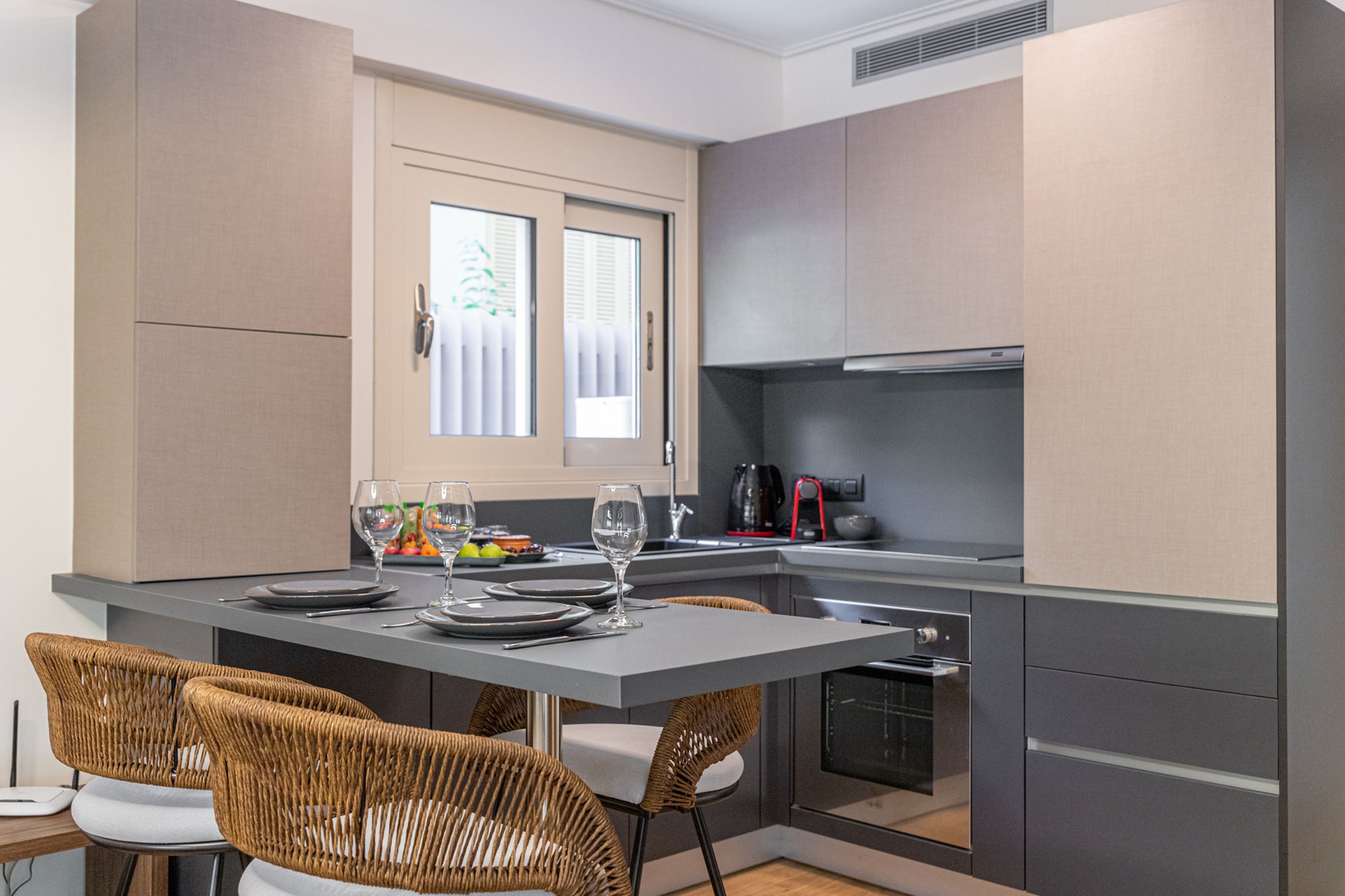 Une cuisine moderne avec des meubles gris, un four encastré et un coin repas avec des chaises en osier et une table dressée pour deux personnes.