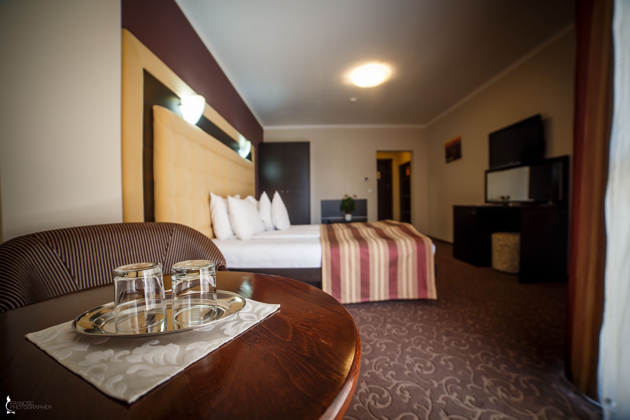 A hotel room with a bed, a desk with a tray of glasses, a television, and a patterned carpet.