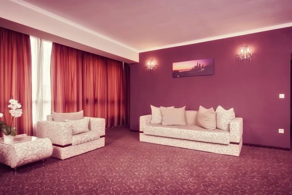 A room with a patterned carpet, two sofas, a small table with a vase of flowers, and a large window with red curtains.