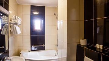 Un baño moderno con una bañera, un inodoro y un lavamanos. Hay toallas apiladas en un perchero y un rollo de papel higiénico en el suelo.
