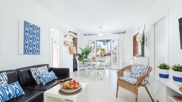 A modern living room with a black leather sofa, a wicker chair, a white coffee table with a bowl of fruit, and a dining area with white chairs and a table.