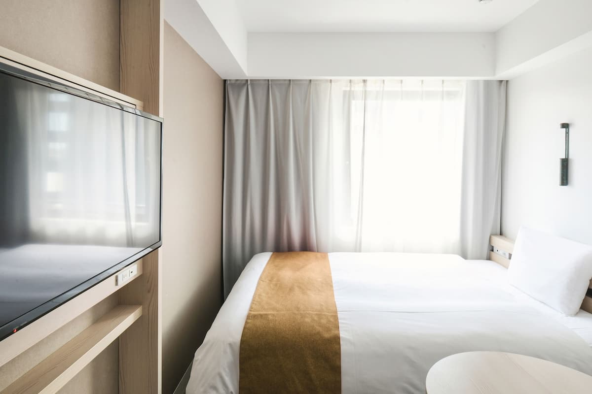 A hotel room with a bed, a TV, a bedside table, and a window with curtains.