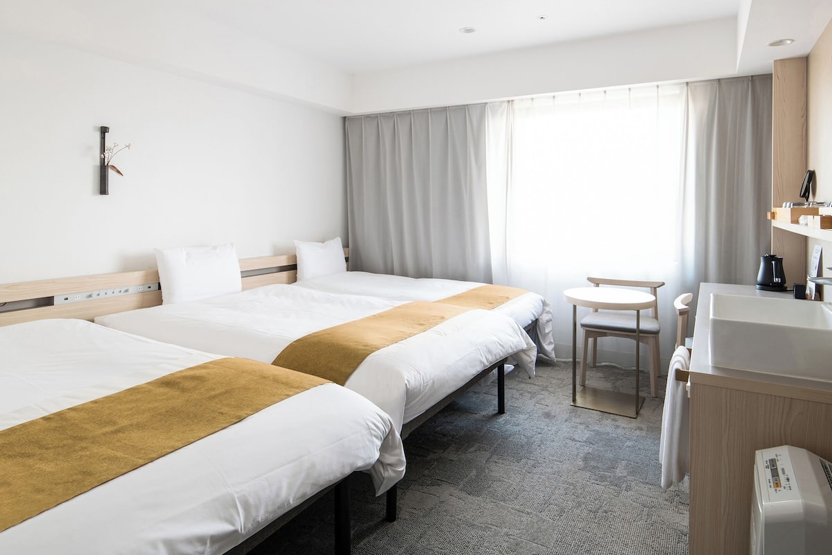 A hotel room with two beds, a small desk, a chair, and a window with curtains.