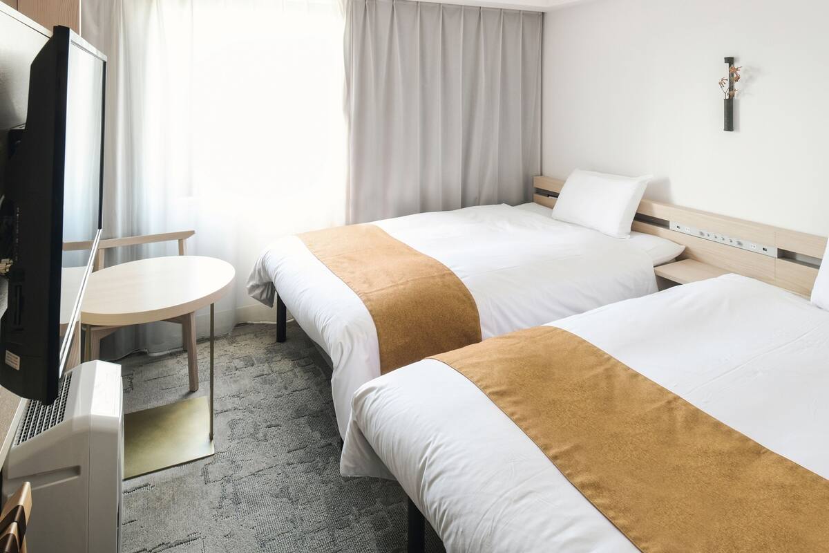 A hotel room with two beds, a desk, a TV, and a window with curtains.