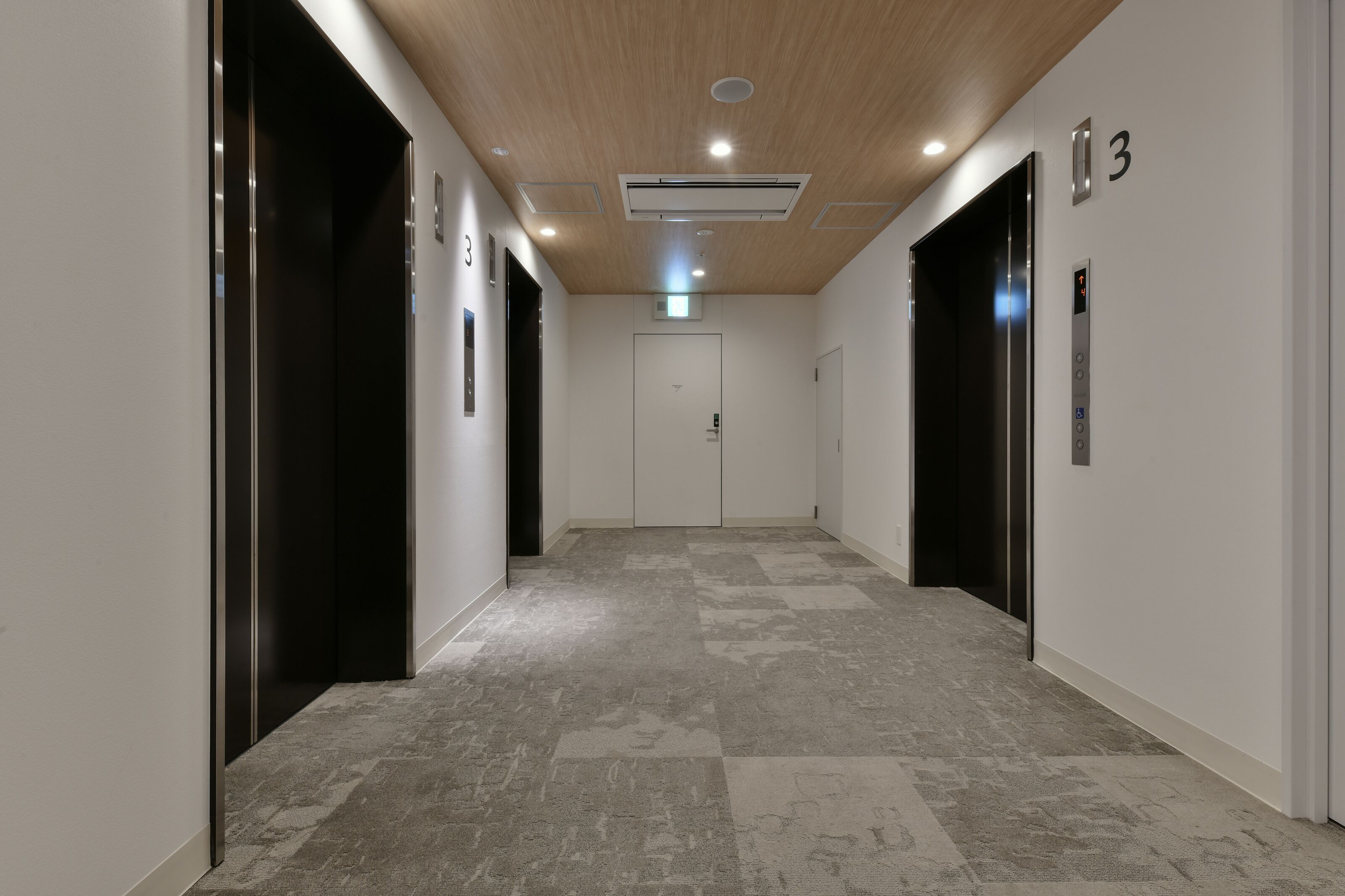 A modern hallway with two elevators, a light-colored carpet, and a wooden ceiling.