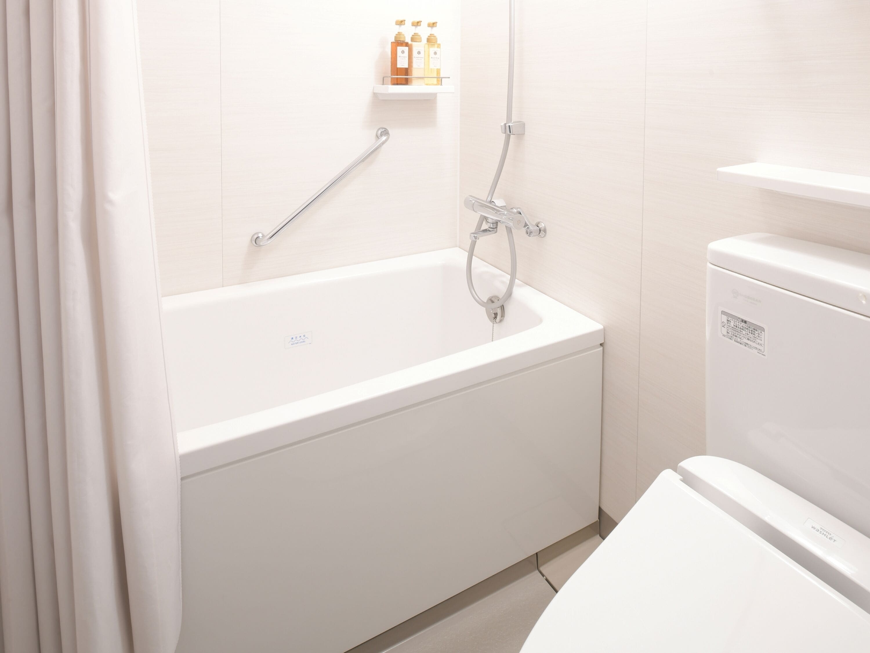 Ein modernes Badezimmer mit einer weißen Badewanne, einer Toilette und einer Duschkabine. Über der Badewanne befindet sich ein Ablagefach mit Pflegeprodukten.
