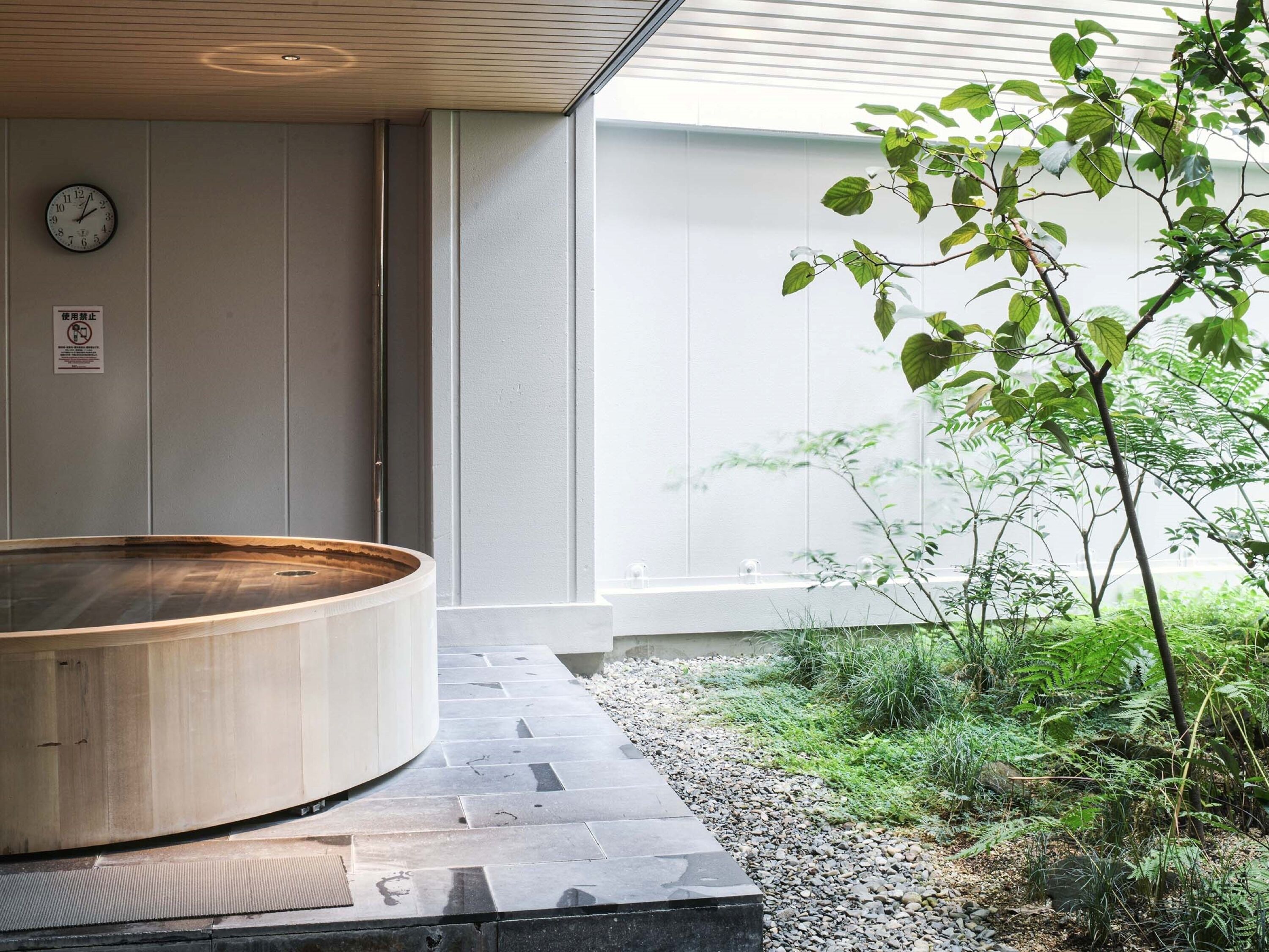 Una vasca idromassaggio in legno in una zona spa, con un vialetto in pietra, circondata dal verde e vista su un edificio con l'orologio.
