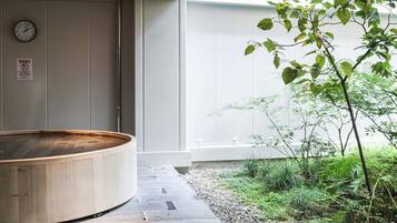 Una vasca idromassaggio in legno in una zona spa, con un vialetto in pietra, circondata dal verde e vista su un edificio con l'orologio.