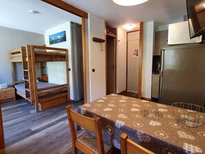 A compact room with a bunk bed, a dining table with a patterned tablecloth, a refrigerator, and a television.