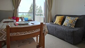A room with a dining table set for two, a sofa with patterned cushions, and a view through a large window. - Southwest cabin studio 4/6 pers near slopes in Chamrousse (Chamrousse)