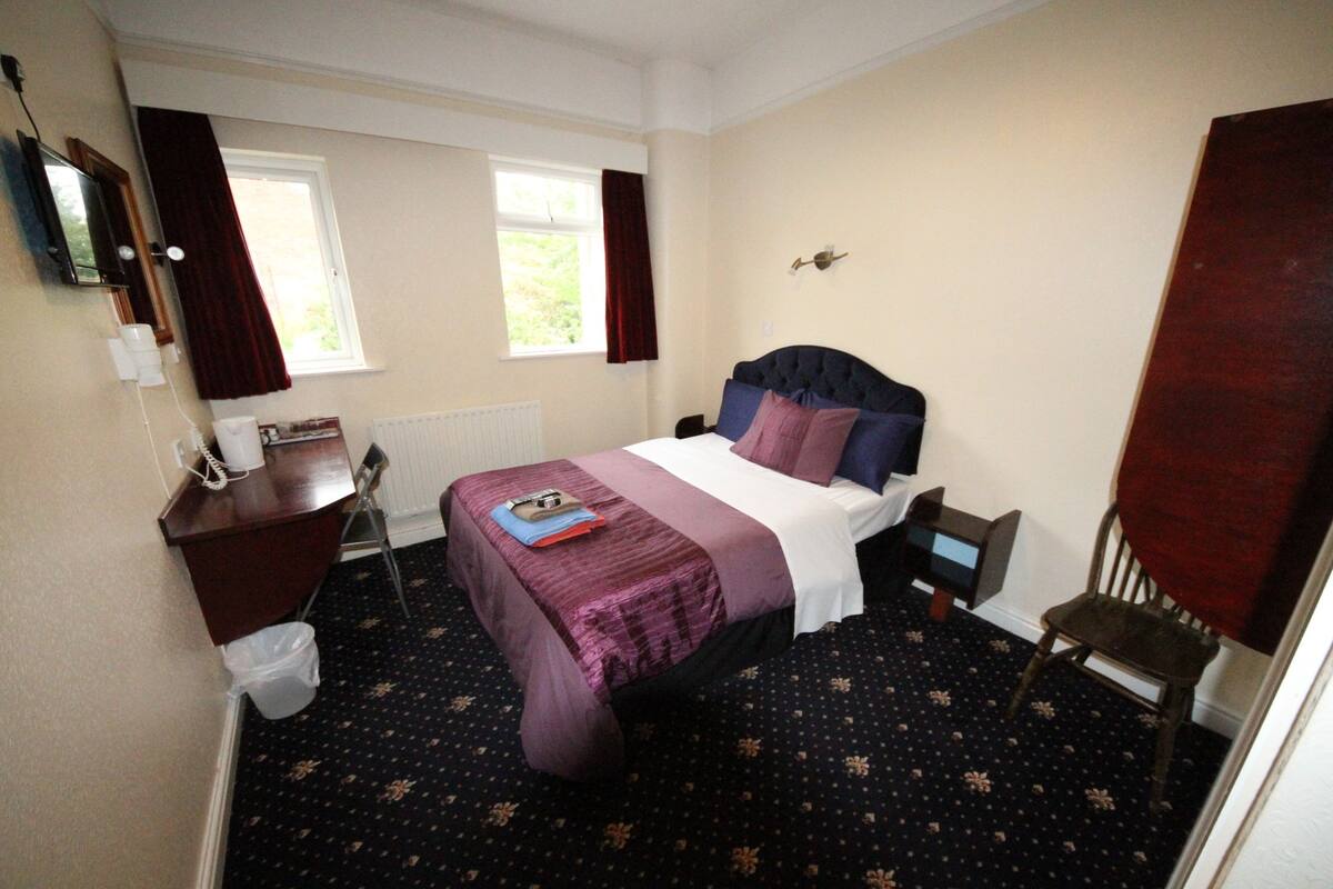 A bedroom with a bed, a desk, a chair, a radiator, a window, and a door.