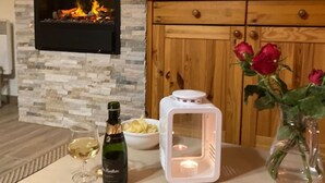 Une pièce avec une cheminée en pierre, un meuble en bois, une bouteille de vin, un bol de chips, une bougie et un vase rempli de roses.