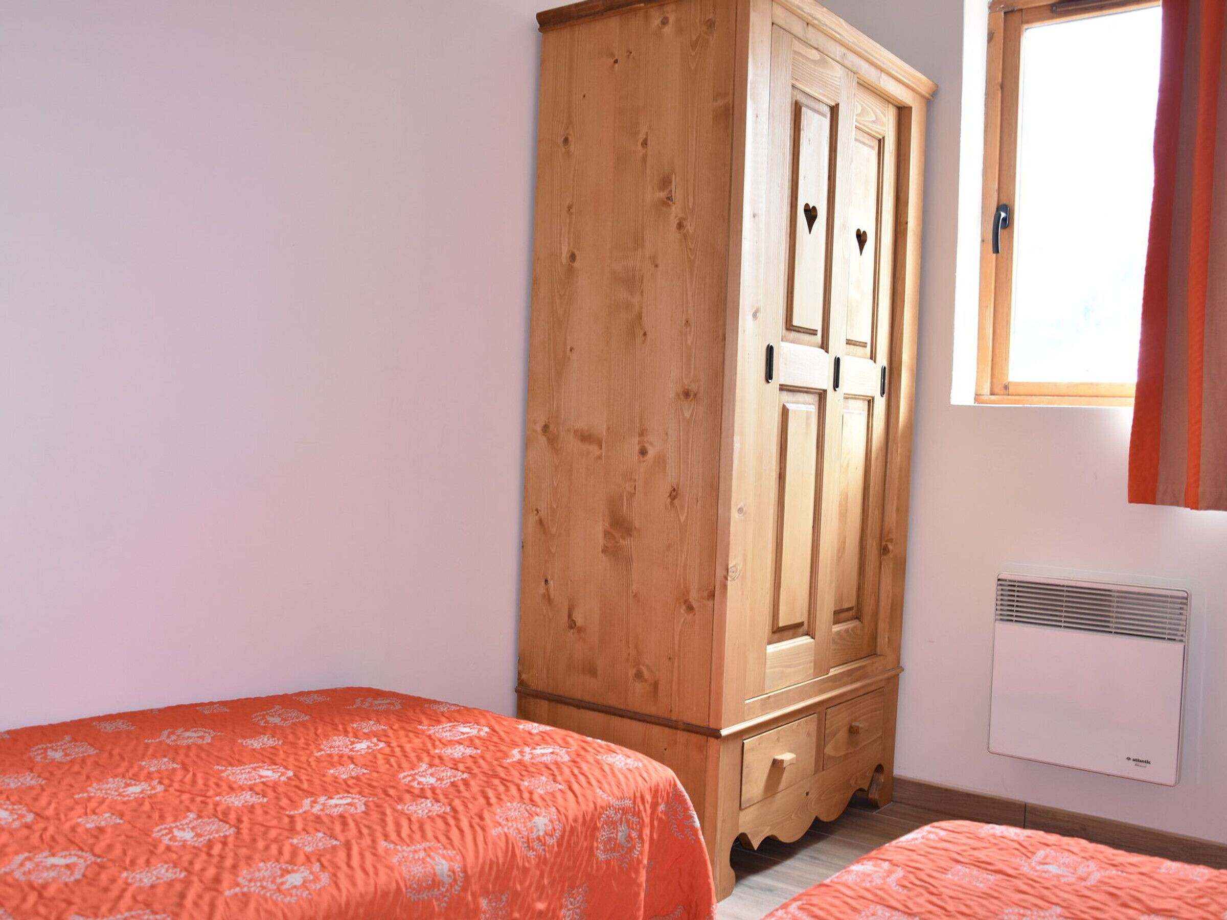 A bedroom with two single beds, a wooden wardrobe, a window with curtains, and a wall-mounted air conditioner.