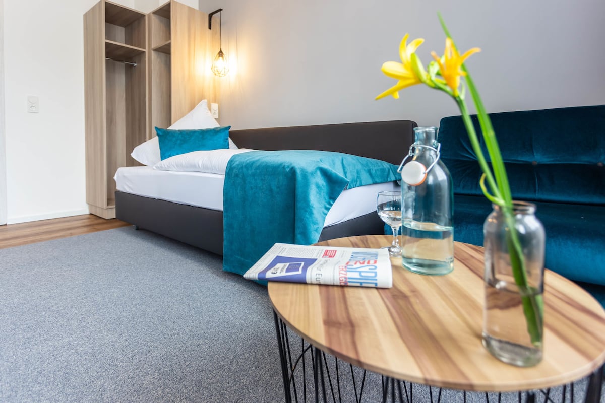 Una habitació d'hotel moderna amb un llit, una taula de fusta amb una ampolla de vidre i un ram de flors, una revista i un sofà blau.