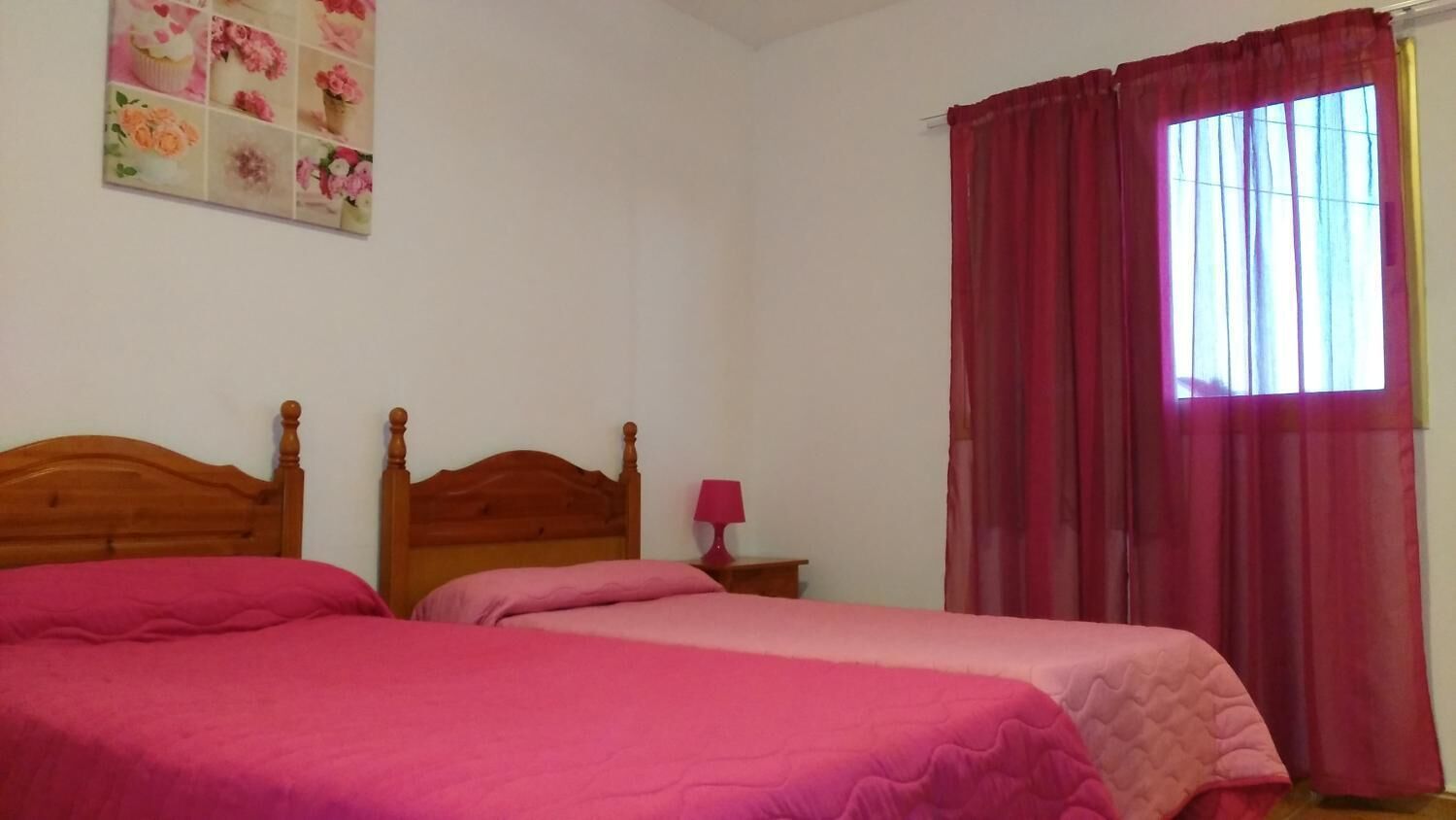 A bedroom with two beds, a wooden headboard, a nightstand with a lamp, and a window with red curtains.