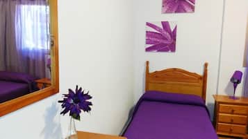 Quarto com cama de madeira, roupa de cama roxa, um espelho, um vaso com uma flor roxa e duas pinturas penduradas na parede.
