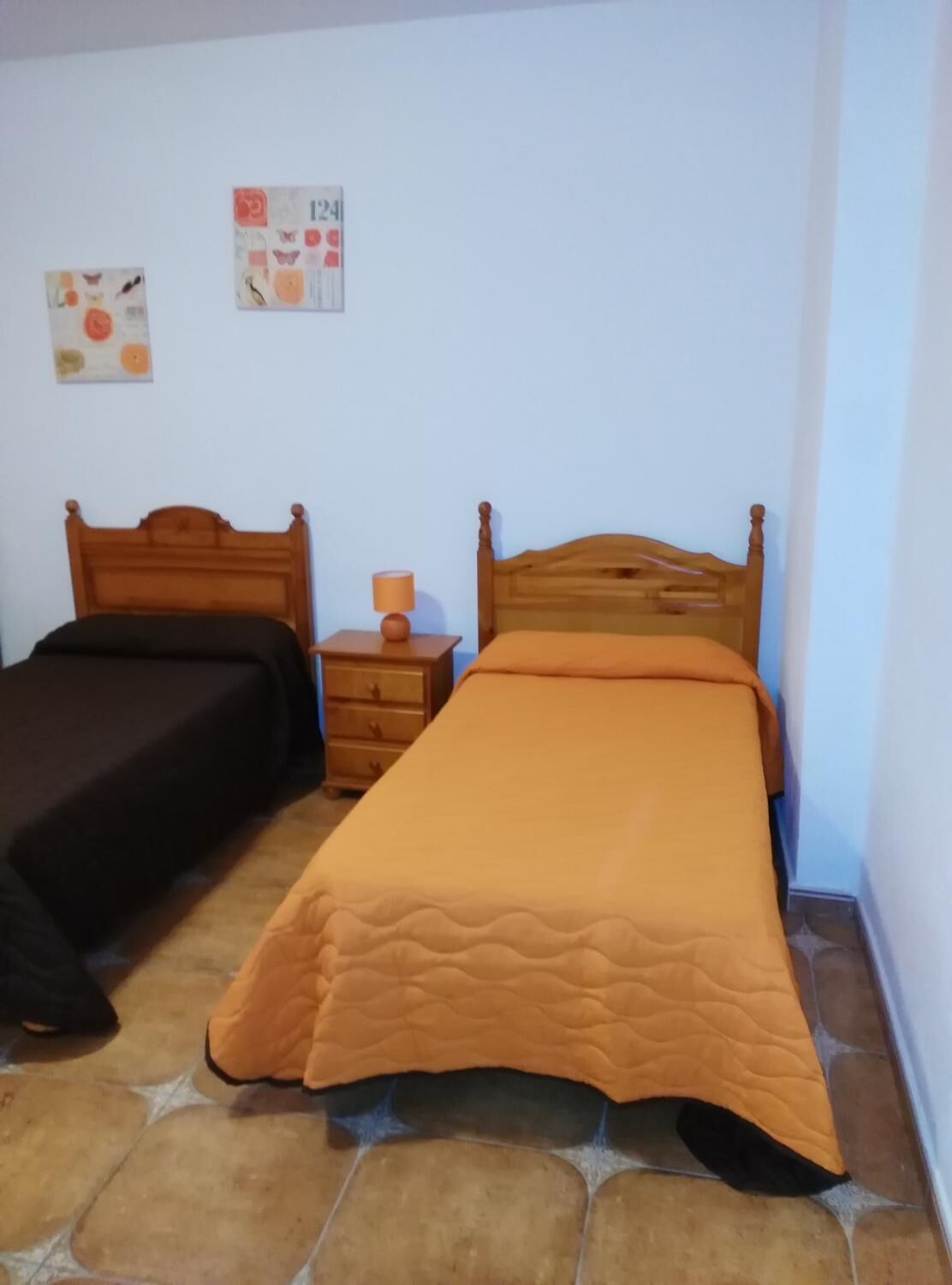 Un dormitorio con dos camas de madera, una mesita de noche con lámpara y una pared con dos cuadros enmarcados.