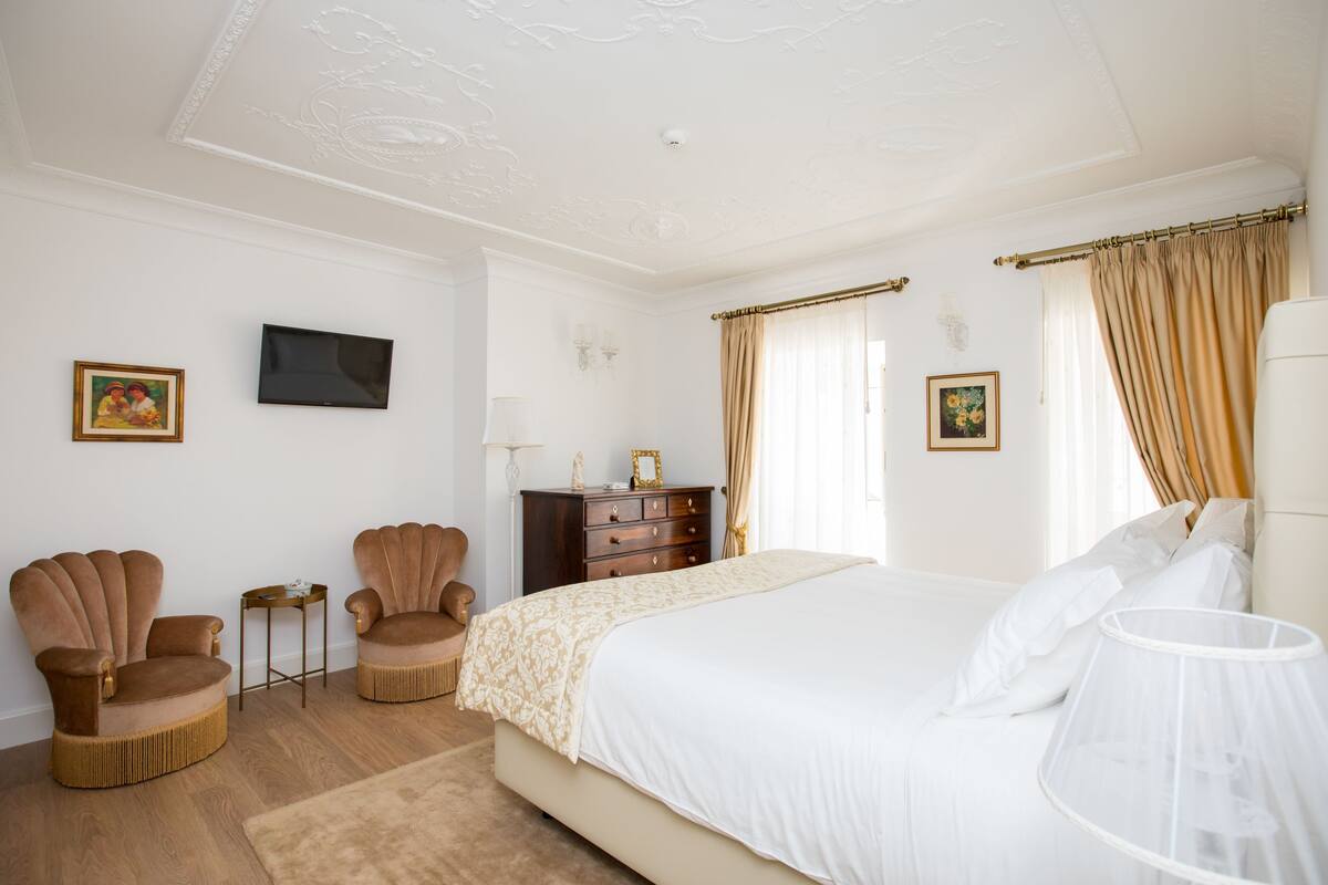 A bedroom with a large bed, two armchairs, a dresser, and a television on the wall.