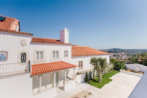 Un bâtiment blanc avec des toits recouverts de tuiles rouges, un balcon doté d’une balustrade et une vue sur une ville au loin.