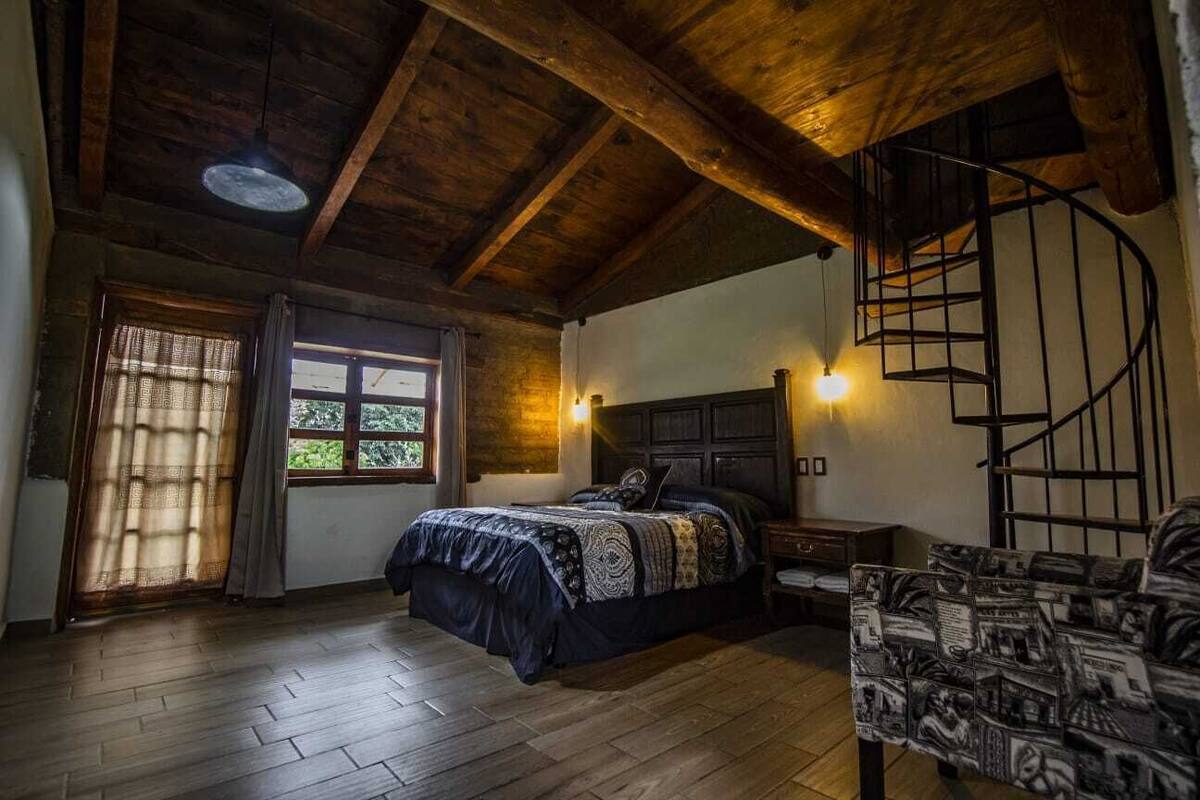 Een slaapkamer met een houten plafond, een spiltrap, een bed met behangpatroon, een nachtkastje en een stoel met krantenprints.