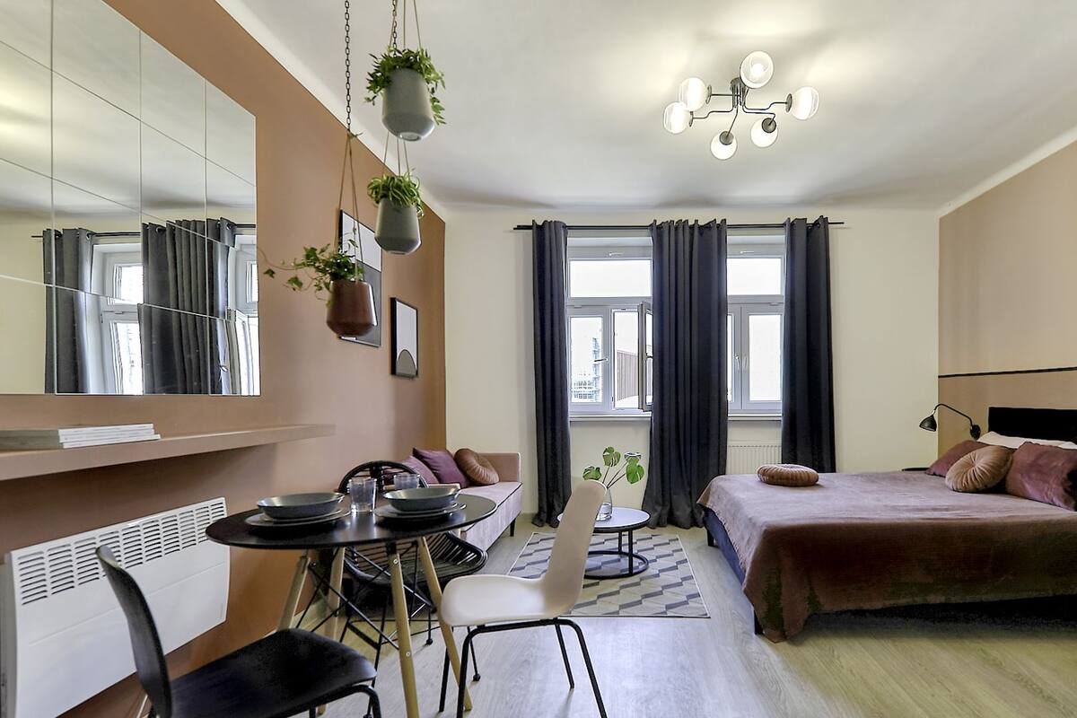 A modern hotel room with a dining area, a bed with a headboard, a large mirror, and a view of the city.
