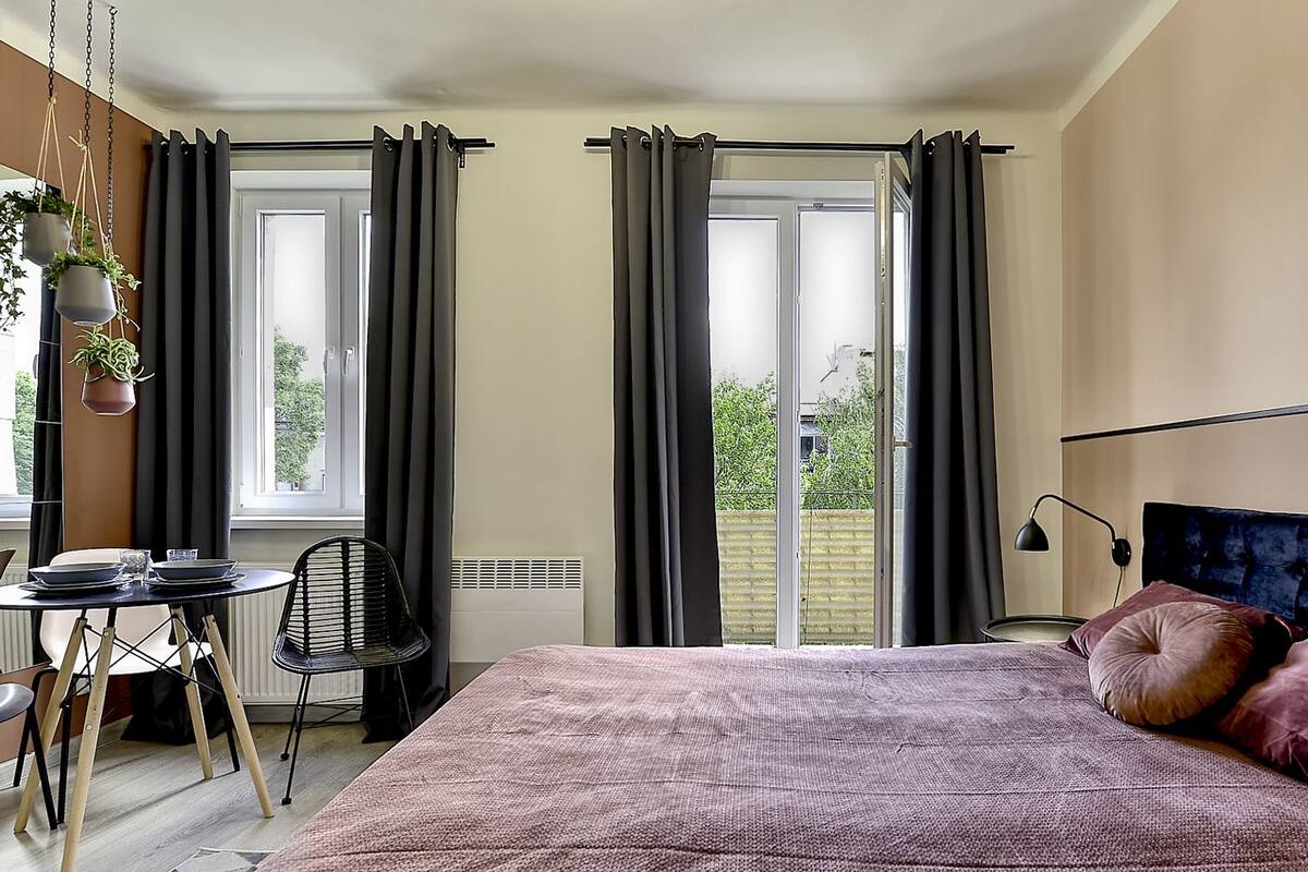 A bedroom with a bed, a desk, and a chair. There are two windows with curtains, and a view of the outside.