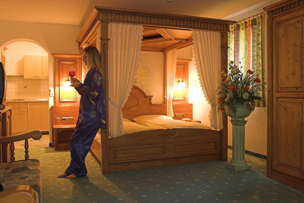 A four-poster bed with a canopy, a wooden nightstand with a lamp, a wooden wardrobe, and a potted plant.
