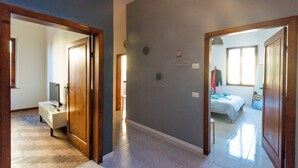 A hallway with a wooden floor, a white bench, and a door leading to a bedroom with a bed and a window. - Your home in Trequanda (Trequanda)