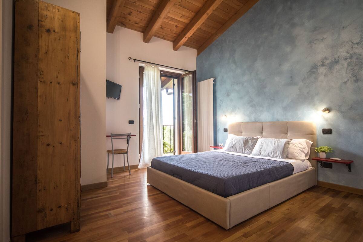 A bedroom with a wooden floor, a bed with a headboard, a nightstand, a small table, and a window with curtains.