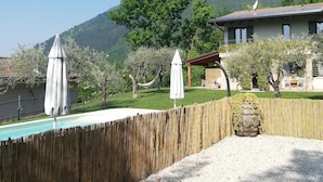 A pool area with a wooden fence, two white umbrellas, and a house with a mountain in the background. - B&B Al Sentiero (Caprino Veronese)
