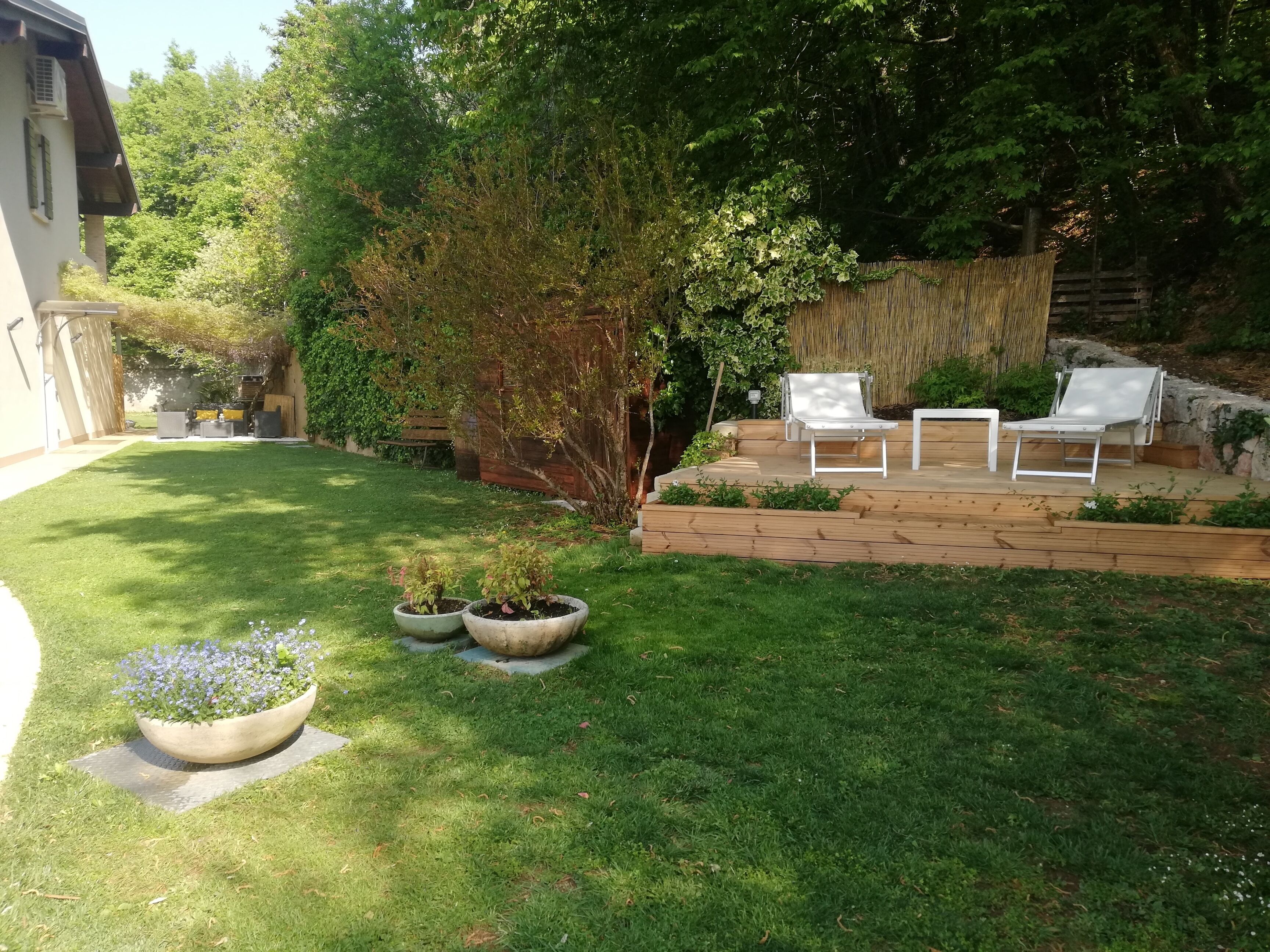 Un patio trasero con césped, una terraza de madera con sillas blancas y un pequeño jardín con plantas en macetas.