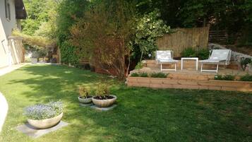 Un patio trasero con césped, una terraza de madera con sillas blancas y un pequeño jardín con plantas en macetas.