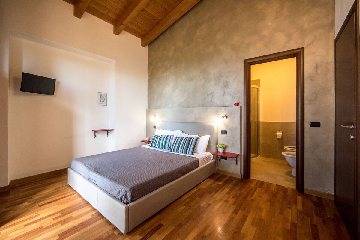A bedroom with a wooden floor, a bed with a gray blanket, a TV mounted on the wall, and a bathroom visible through an open door.