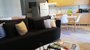 A modern living room with a black sofa, a coffee table with magazines, and a kitchen area with wooden cabinets and a microwave.