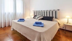 Un lit bien fait, recouvert d’une couverture beige, avec deux oreillers et deux serviettes bleues pliées. Il y a deux tables de chevet avec des lampes, et une fenêtre avec des rideaux.