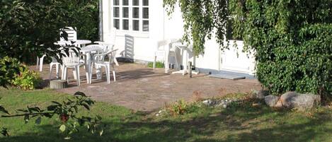 Een wit gebouw met een pannendak, een klein terras met witte plastic stoelen, en een grote boom op de voorgrond.