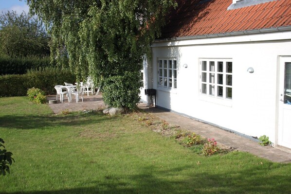 A white building with a tiled roof, a small garden with grass and a few plants, and white plastic chairs arranged outside. - Spangsgaard bed and breakfast (Odense)