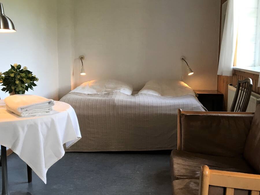 A neatly made bed with a white pillow and a folded white towel on a table beside it. There is a small potted plant on the table.