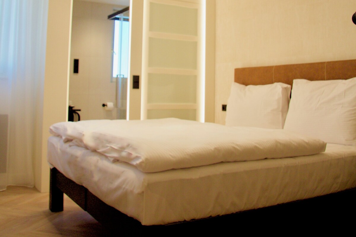Quarto de hotel moderno com uma cama grande, roupeiro embutido e luminária circular no teto.